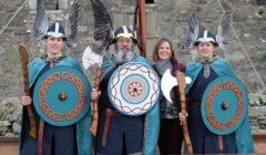 Three people dressed as Vikings with shields and axes stand next to a smiling woman in modern clothes, in front of a stone building.