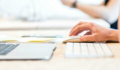 Person's hand using a computer mouse next to a keyboard and an open laptop on a desk.