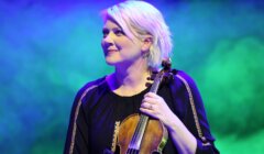 A person with short blond hair holds a violin on stage, wearing a black top with decorative trim, set against a colorful, blurred background.