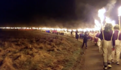 A nighttime procession with people carrying lit torches, walking along a path through a grassy area.