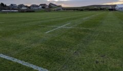 A grassy sports field with white boundary lines, set against a backdrop of houses and a cloudy sky.