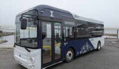 A modern white and navy blue city bus labeled "HELLO" and "CIAO" is parked on a paved surface near a body of water on a cloudy day.