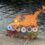 A painted cardboard boat with paper decorations is on fire at the edge of a body of water.