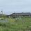 A low, single-story building sits behind a grassy field with goalposts, a sign, and a parked car under an overcast sky.