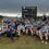 A rugby team poses in front of a scoreboard reading "Deeside Rugby Home 24 Visitors 63" outdoors on a cloudy day.