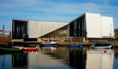 A modern angular building with large glass windows sits by a harbor, with several colorful small boats floating on the calm water in front.