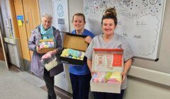 Three women stand in a hallway holding boxes filled with handmade baby clothes. Two are in scrubs, one in a coat. Behind them is a whiteboard with baby-themed drawings and notes.