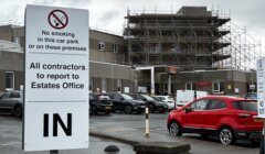Sign at a car park entrance reads "No smoking in this car park or on these premises. All contractors to report to Estates Office. IN." Scaffolding on a building in the background.