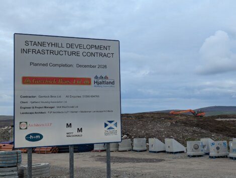 Sign for Staneyhill Development Infrastructure Contract with contractor details in foreground; construction site and machinery visible in the background under a cloudy sky.