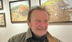 An older man wearing a coat stands in front of landscape paintings displayed on a gallery wall.