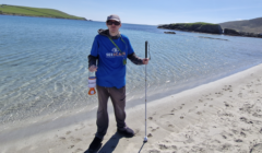 A person wearing sunglasses and a blue "SeeScape" shirt stands on a sandy beach, holding a litter picker and bottle, with clear water and hills in the background.
