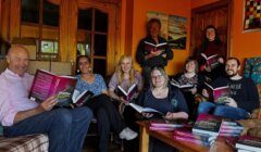 A group of seven people sit and stand in a cozy, book-filled room, each holding an open book and smiling at the camera.