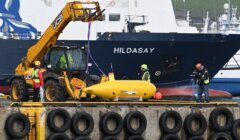 A yellow underwater vehicle is lifted by a crane on a dock, with several workers in safety gear, and a large ship named HILDASAY in the background.