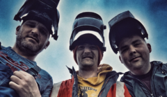 Three people wearing welding helmets stand outdoors, looking down at the camera. The sky is cloudy and the overall tone of the image is cool and slightly dark.