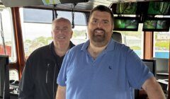 Two men stand inside a control room with multiple monitors and windows, with one man wearing a black jacket and the other in a blue polo shirt.