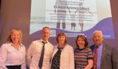 Five people stand in front of a presentation slide about the Viking Genes Shetland Community Screening Project fundraising drive.