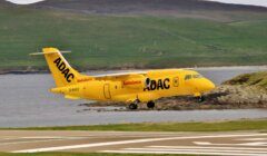 A yellow ADAC ambulance plane is landing on a runway near a body of water, with green hills in the background.
