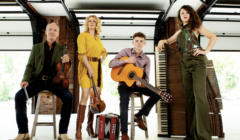 Four musicians pose in a garage with instruments: a violin, guitar, accordion, and an upright piano in the background. Three are standing and one is seated, all looking at the camera.