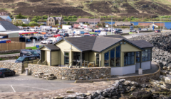 A modern building with large windows is situated next to a stone wall, overlooking a parking lot with multiple vehicles and a hillside in the background.