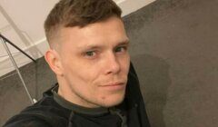 A man with short light brown hair and a trimmed beard wears a dark shirt and sits on a carpeted floor, looking at the camera.