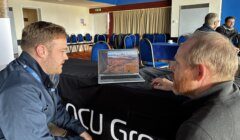 Two men sit at a table, looking at a laptop displaying an aerial landscape photo, in a room with blue chairs and people in the background.