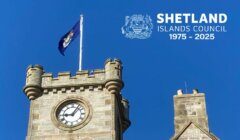 A stone clock tower with a flag on top against a blue sky. Text reads "Shetland Islands Council 1975-2025" with a crest.