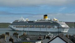 A large cruise ship is anchored near the shore, with rooftops of houses and docks visible in the foreground under a partly cloudy sky.