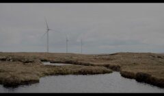 Three wind turbines stand in a grassy, open landscape near a body of water under an overcast sky.