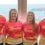 Four people stand indoors in front of a window with a coastal view, all wearing matching red and yellow Brain Tumour Research shirts, facing the camera and smiling.