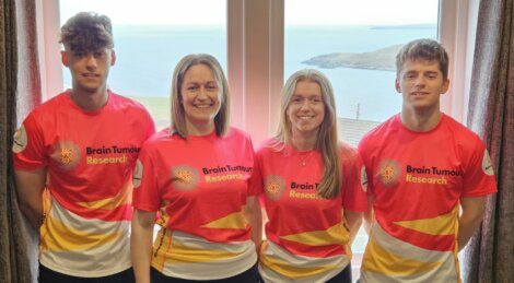 Four people stand indoors in front of a window with a coastal view, all wearing matching red and yellow Brain Tumour Research shirts, facing the camera and smiling.