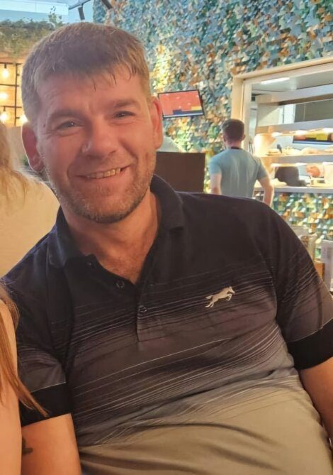 A man with short brown hair and a beard is smiling while sitting indoors at a restaurant with a kitchen visible in the background.