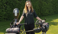 A woman stands outdoors holding a loaded touring bicycle with panniers and a cello case attached to the rear.