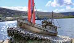 A metal sculpture of a fishing boat sits on a rocky base near the water, with a red flag and coastal landscape in the background.