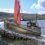 A metal sculpture of a fishing boat sits on a rocky base near the water, with a red flag and coastal landscape in the background.