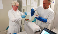 Two scientists in lab coats and blue gloves handle sample trays at a laboratory workstation, with a laptop displaying a spreadsheet nearby.