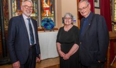 Three people stand indoors in front of a table with a blue glass sculpture, with stained glass windows in the background.