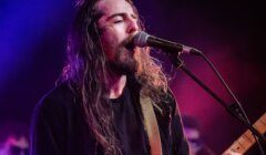 A long-haired musician sings into a microphone and plays electric guitar on stage under colorful lighting.