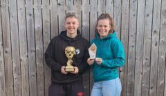 Two young adults stand in front of a wooden fence, each holding a trophy and smiling at the camera.