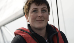 A person with short brown hair wearing a red life jacket stands on a boat, with part of the sail and rigging visible in the background.