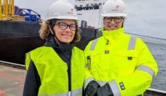Two people wearing hard hats, safety glasses, and high-visibility jackets stand on a dock with a ship and water in the background.