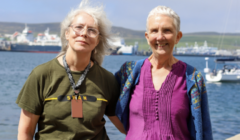 Two older women stand side by side outdoors near a body of water with boats and ships in the background on a sunny day.