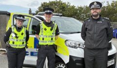 Three police officers in uniform stand in front of a police vehicle parked outdoors on a cloudy day.