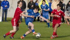 Three teenage soccer players compete for the ball on a grassy field, with two in red uniforms defending against one in blue. Spectators watch from the sidelines.