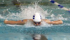 A swimmer wearing a swim cap and goggles performs the butterfly stroke in a pool, creating a splash of water behind them.
