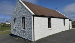 A small white stone building with a brown tiled roof, three windows, and an asphalt area, set in a rural area under a blue sky.