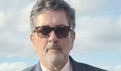 A man with gray hair and a beard wearing sunglasses, a suit, and a tie stands outdoors against a backdrop of cloudy sky.