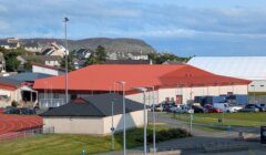 A sports complex with a red roof, parking lot, outdoor running track, and surrounding buildings, set against a hilly landscape.