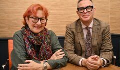 Two people with glasses, one wearing a scarf and green top, the other in a checked suit and tie, sit side by side at a wooden table against a beige wall.