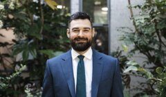 A bearded man in a blue suit and teal tie stands in front of leafy green plants, smiling at the camera.