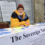 An older man in a yellow jacket and knit hat sits at an outdoor table promoting the Sovereign Nation of Shetland, with informational posters and papers displayed.